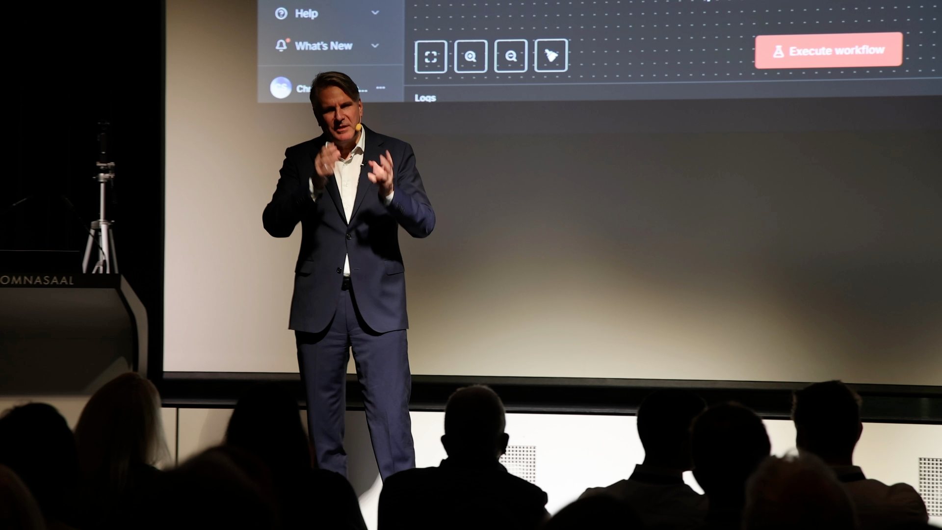 Prof. Dr. Christian Stummeyer auf der Bühne in Rankweil, hinter ihm auf der Leinwand ein KI-Agenten-Workflow